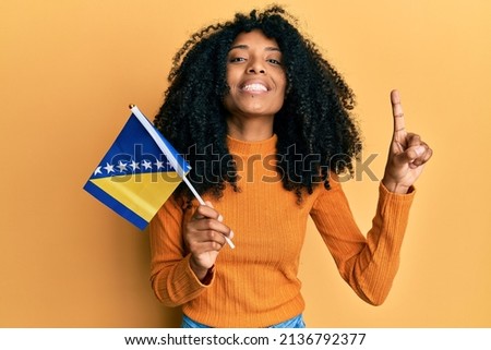 African american woman with afro hair holding bosnia herzegovina flag smiling with an idea or question pointing finger with happy face, number one 