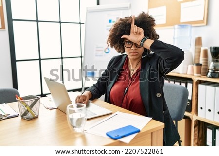 African american woman with afro hair working at the office wearing operator headset making fun of people with fingers on forehead doing loser gesture mocking and insulting. 
