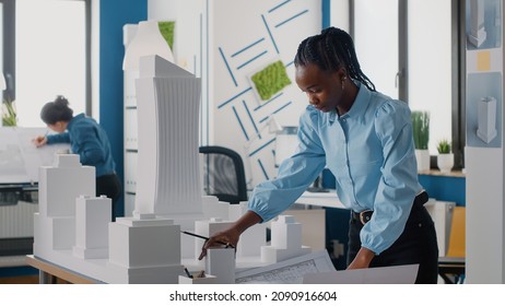 African american woman analyzing blueprints plan and building model to work on architecture. Architect working with maquette and layout print to design urban construction for development.