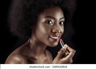 african american woman applying lipstick and looking at camera isolated on black