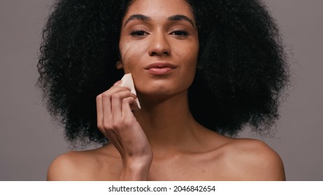 african american woman applying makeup foundation with sponge isolated on grey