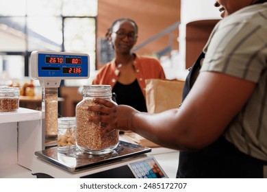 African American woman in an apron assists a customer, weighing locally sourced grains on scale at fresh and organic grocery store. Local merchant measuring jar filled with bio food product. - Powered by Shutterstock