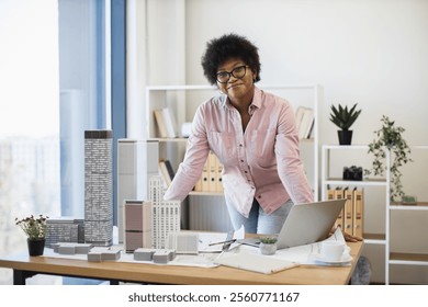African American woman architect in 30s develops project with skyscraper model on desk, wearing glasses.