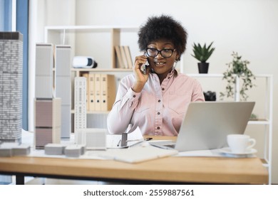 African American woman architect in modern office discussing city project on smartphone, surrounded by building models. Professional setting conveys creativity, architecture, communication.