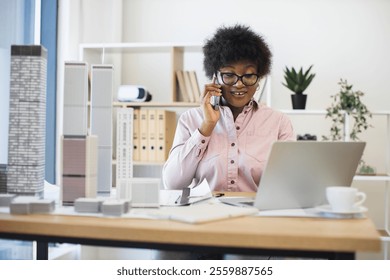 African American woman architect in modern office discussing city project on smartphone, surrounded by building models. Professional setting conveys creativity, architecture, communication.
