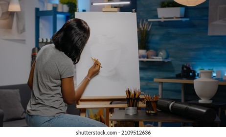 African American Woman With Artist Occupation Drawing Vase On White Canvas Using Pencil While Sitting In Creativity Studio. Black Young Artist Working On Art Masterpiece As Artistic Hobby