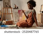 African American woman artist wearing overalls, working on canvas on studio floor, holding brush and sitting with focused expression, surrounded by art supplies and paintings