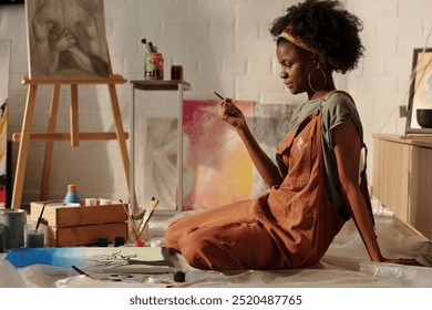 African American woman artist wearing overalls, working on canvas on studio floor, holding brush and sitting with focused expression, surrounded by art supplies and paintings - Powered by Shutterstock