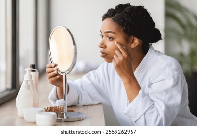 African American woman in a bathrobe focuses intently on her reflection while applying skincare products in a well-lit room. Natural light enhances the serene atmosphere of her morning ritual. - Powered by Shutterstock