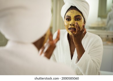 African American Woman In Bathroom Wearing Towel On Head, Looking In Mirror And Applying Face Mask. Health, Beauty And Wellbeing, Spending Quality Time At Home.