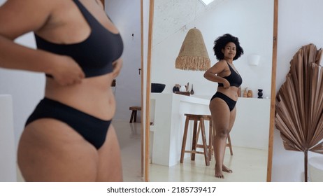 An African American Woman In Black Lingerie Looks At Her Curvaceous Figure In Front Of A Mirror And She Likes Her Body. Acceptance Of Obesity By A Woman. The Concept Of Body Positivity.