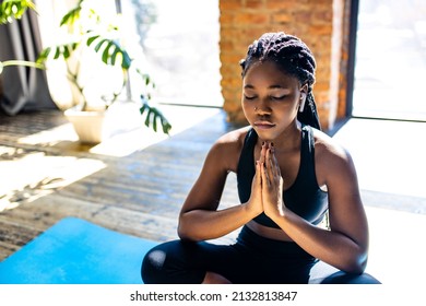 african american woman in black sporty bra is sitting lotus pose on yoga mat floor closed eyes and listening an audio meditate mantra hands namaste
