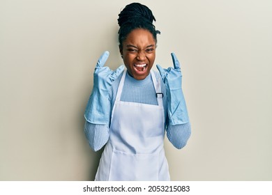 African american woman with braided hair wearing cleaner apron and gloves shouting with crazy expression doing rock symbol with hands up. music star. heavy concept. 