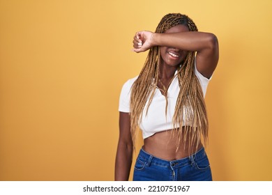 African American Woman With Braided Hair Standing Over Yellow Background Covering Eyes With Arm Smiling Cheerful And Funny. Blind Concept. 