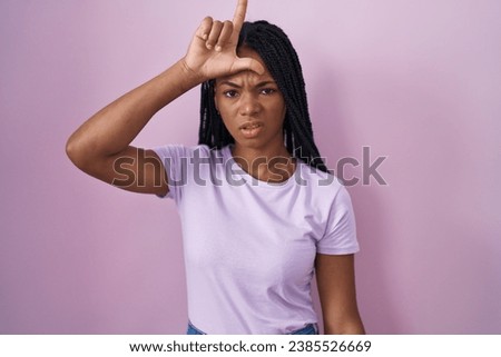 African american woman with braids standing over pink background making fun of people with fingers on forehead doing loser gesture mocking and insulting. 