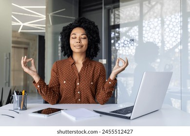 African American woman in a brown polka dot shirt meditating with eyes closed in front of a laptop at her modern office space, promoting wellness and mindfulness at workplace. - Powered by Shutterstock