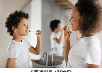 African American woman is brushing her teeth next to a little boy. They are standing in front of a bathroom sink. The woman holds a toothbrush while the little boy watches intently. - Powered by Shutterstock