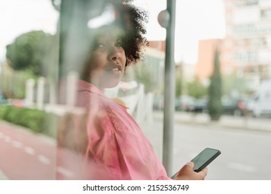 african american woman with bulky curly hair is sitting inside of the outdoor bus stop and using her smartphone, selective focus