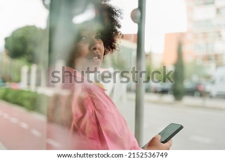african american woman with bulky curly hair is sitting inside of the outdoor bus stop and using her smartphone, selective focus