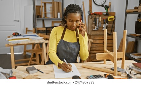 An african american woman carpenter multitask in her workshop, taking notes while talking on the phone.