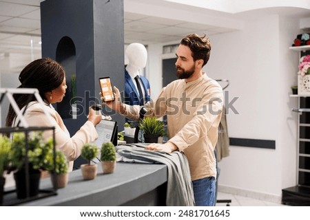 African American woman cashier scanning Black Friday mobile promo code from customer smartphone, using barcode scanner. Young man shopper using coupon to get discount while shopping in clothing store