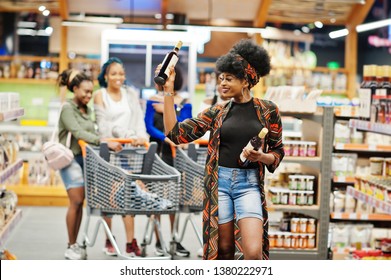 African American Woman Choose Wine At Supermarket Against Her Afro Friends With Shopping Cart.