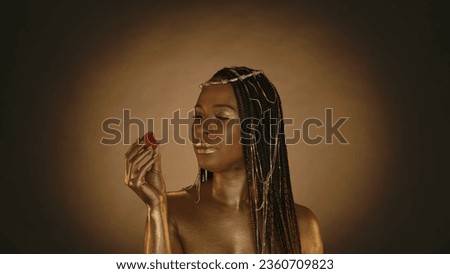 African American woman in Cleopatra style in the studio on brown background with circular light. A woman with golden skin and jewelry on her head holds juicy ripe strawberries. Fashion art design.