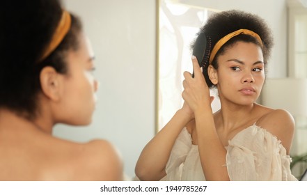 African american woman combing her hair with hairbrush, makes hairstyle in front of the mirror at home. Getting ready in morning. - Powered by Shutterstock