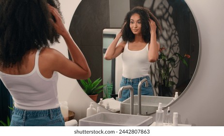 African American woman come in home bathroom looking at mirror reflection beautiful pretty ethnic girl female lady fix curls check curly hair doing hairdo hairstyle beauty routine shampoo go away - Powered by Shutterstock