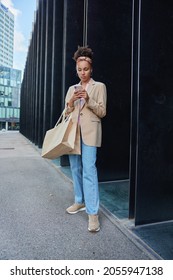 African American Woman Creats Review About Black Friday Sales On Website Uses Cellular Phone Poses With Paper Shopping Bags Outdoors Bought Fashionable Clothes In Mall Concentrated At Screen