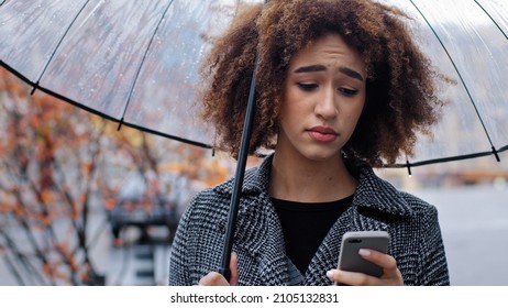 African American Woman Curly Girl With Transparent Umbrella Stands On Autumn Street In Rain Looks At Mobile Phone Upset Sadness Worrying About Low Battery Problems With Internet Bad News Gets Rejected