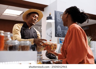 African American woman discusses eco friendly shopping with the shopkeeper at the checkout counter. A female customer giving the jar of pasta to the male vendor at the cashier desk. - Powered by Shutterstock