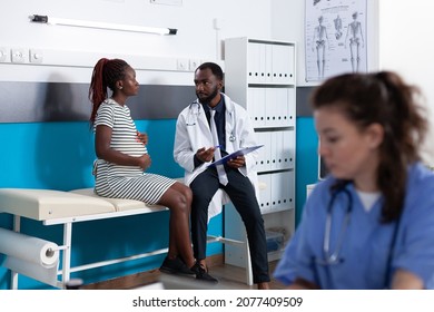 African American Woman And Doctor Talking About Pregnancy At Medical Appointment In Office. Patient Expecting Child And Having Discussion With Physician To Check Healthcare. Pregnant Adult