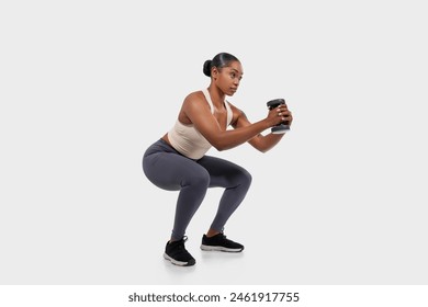 African American woman is doing a squat exercise while holding a dumbbell in her hand. She is lowering her body down by bending her knees and hips, then pushing back up to the starting position - Powered by Shutterstock