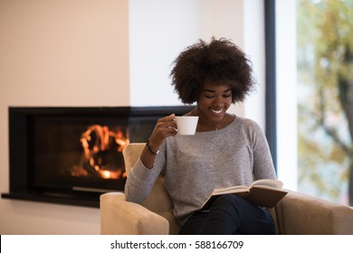 African American Woman Drinking Cup Of Coffee Reading Book At Fireplace. Young Black Girl With Hot Beverage Relaxing Heating Warming Up. Autumn At Home.