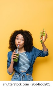 African American Woman Drinking Fresh Smoothie While Holding Hot Dog On Yellow