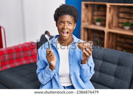 African american woman eating healthy whole grain cereals angry and mad screaming frustrated and furious, shouting with anger looking up. 