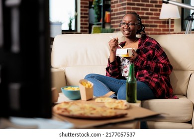 African American Woman Eating Noodles From Takeout Delivery Package And Relaxing On Couch With Movie On Tv. Using Choppsticks To Eat Takeaway Fast Food Meal And Drinking Beer From Bottle.