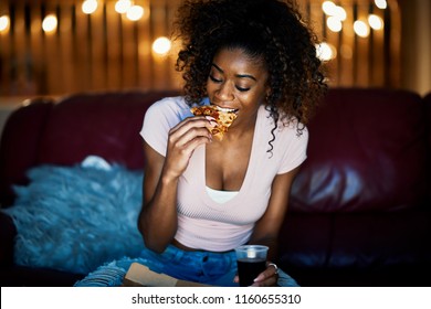 African American Woman Eating Pizza And Drinking Soda Late At Night On Sofa While Watching Tv