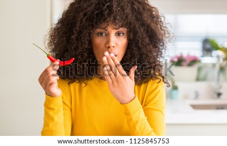 African american woman eating red hot chili pepper cover mouth with hand shocked with shame for mistake, expression of fear, scared in silence, secret concept