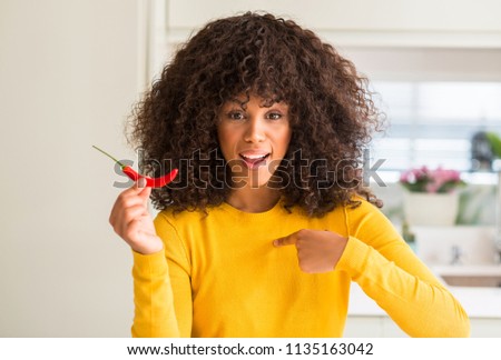 African american woman eating red hot chili pepper with surprise face pointing finger to himself