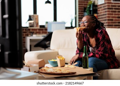 African American Woman Eating Slice Of Pizza And Watching Movie, Relaxing On Couch With Takeaway Food Delivery. Enjoying Fast Food Meal From Takeout Package To Watch Film On Television.