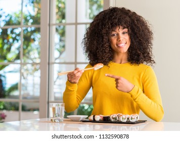 African american woman eating sushi using chopsticks at home very happy pointing with hand and finger