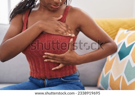 African american woman examining breast with hand sitting on sofa at home