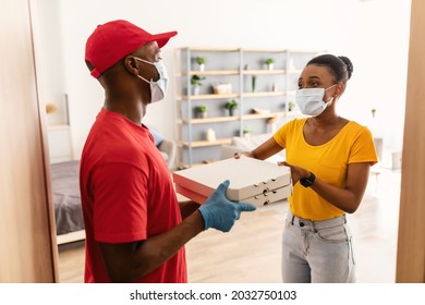 African American Woman In Face Mask Receiving Pizza Boxes From Restaurant Delivery Man Wearing Face Masks Standing At Doors Of Her Home. Courier Service And Food Delivery Concept