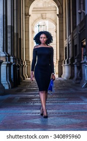 African American Woman Fashion in New York. Wearing long sleeve, slim, off shoulder dress, carrying blue bag, a young lady walking on narrow street, going to work. Filtered look with dark blue tint.