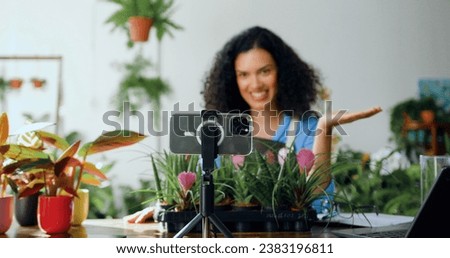 African American woman florist in apron shooting video in flower store using phone. Gardener blogger using phone broadcasting live on smartphone for online shopping in social networks.