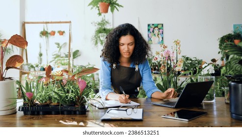 African american woman florist using laptop entrepreneur taking notes wearing apron. Female flower shop manager small business owner working in flower shop, plant store. Organizes logistics and - Powered by Shutterstock