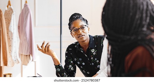 African American woman in glasses, in conversation. African American woman fashion designer discussing, with clothes hanging in the background. Fashion design business with African American women. - Powered by Shutterstock