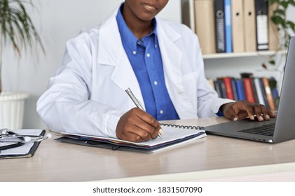 African american woman gp doctor wears white coat using laptop computer writes patient prescription notes records sits at desk. Black professional medic working at workplace in hospital. Close up view